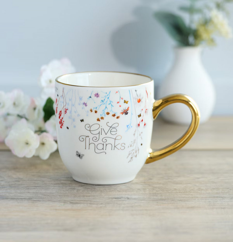 White mug with gold handle and 'Give Thanks' text on a wooden surface with flowers in the background