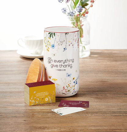 Decorative container with floral design and text on a wooden surface with a cup and vase in the background.