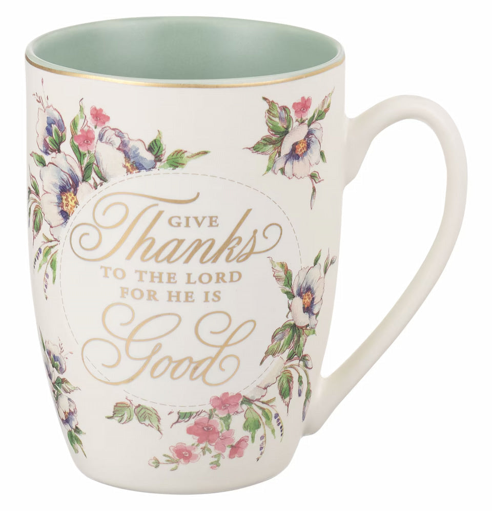 A white ceramic coffee mug with floral patterns in pink and blue colors, and a gold-foiled text that reads 'Give Thanks to the Lord for He is Good' on the front. The back of the mug features the gold-foiled Scripture reference 'Psalm 107:1'.