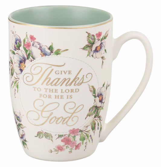 A white ceramic coffee mug with floral patterns in pink and blue colors, and a gold-foiled text that reads 'Give Thanks to the Lord for He is Good' on the front. The back of the mug features the gold-foiled Scripture reference 'Psalm 107:1'.