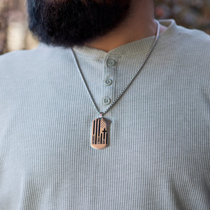 Person wearing a necklace with a dog tag featuring a cross and stripes design.