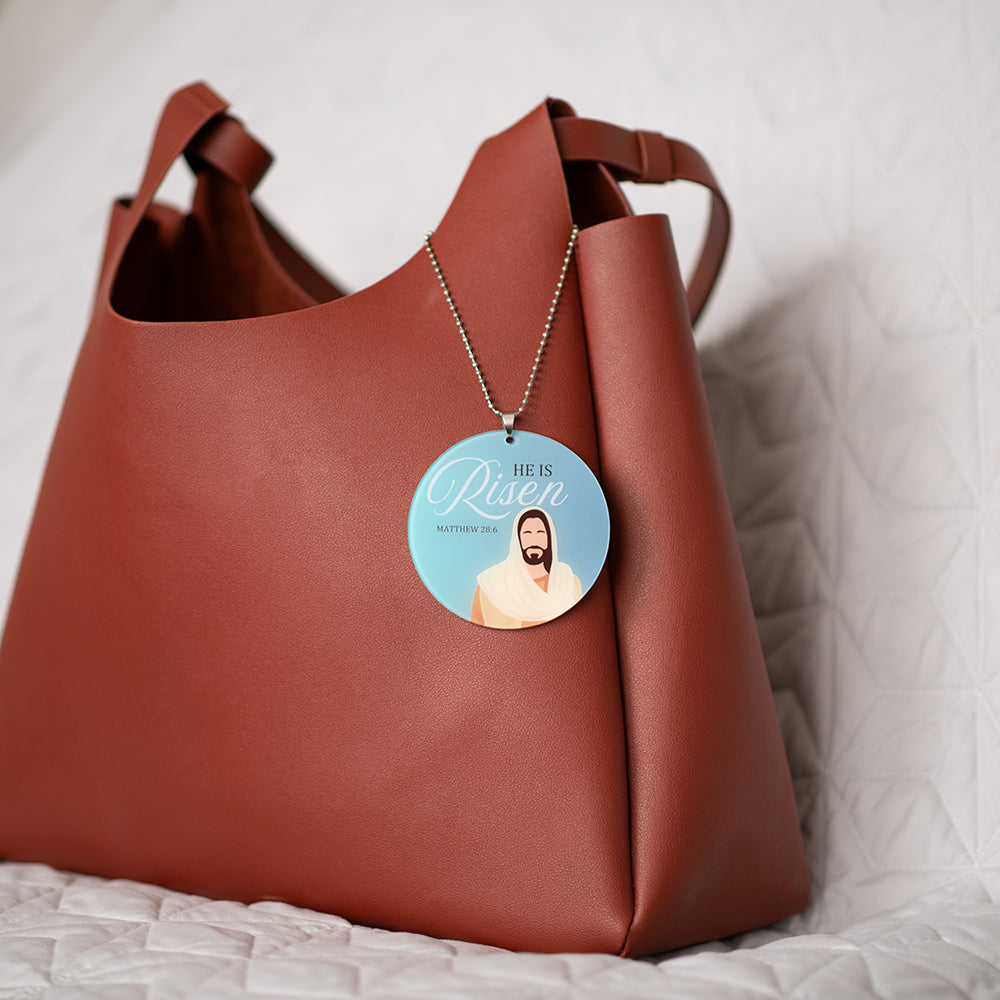 Brown leather handbag with a 'He is Risen' necklace on a white background