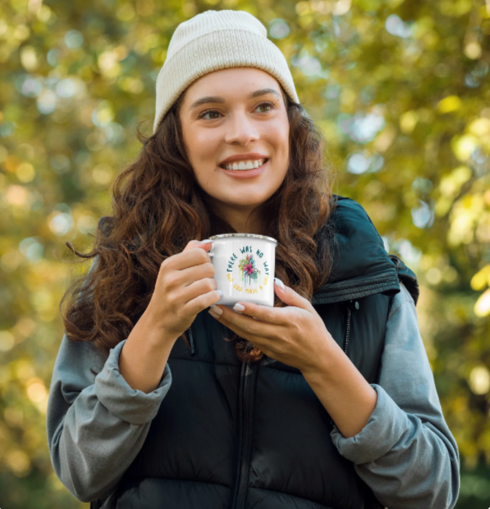 A person wearing a beige beanie and a gray vest is holding a white enamel mug with a floral design.