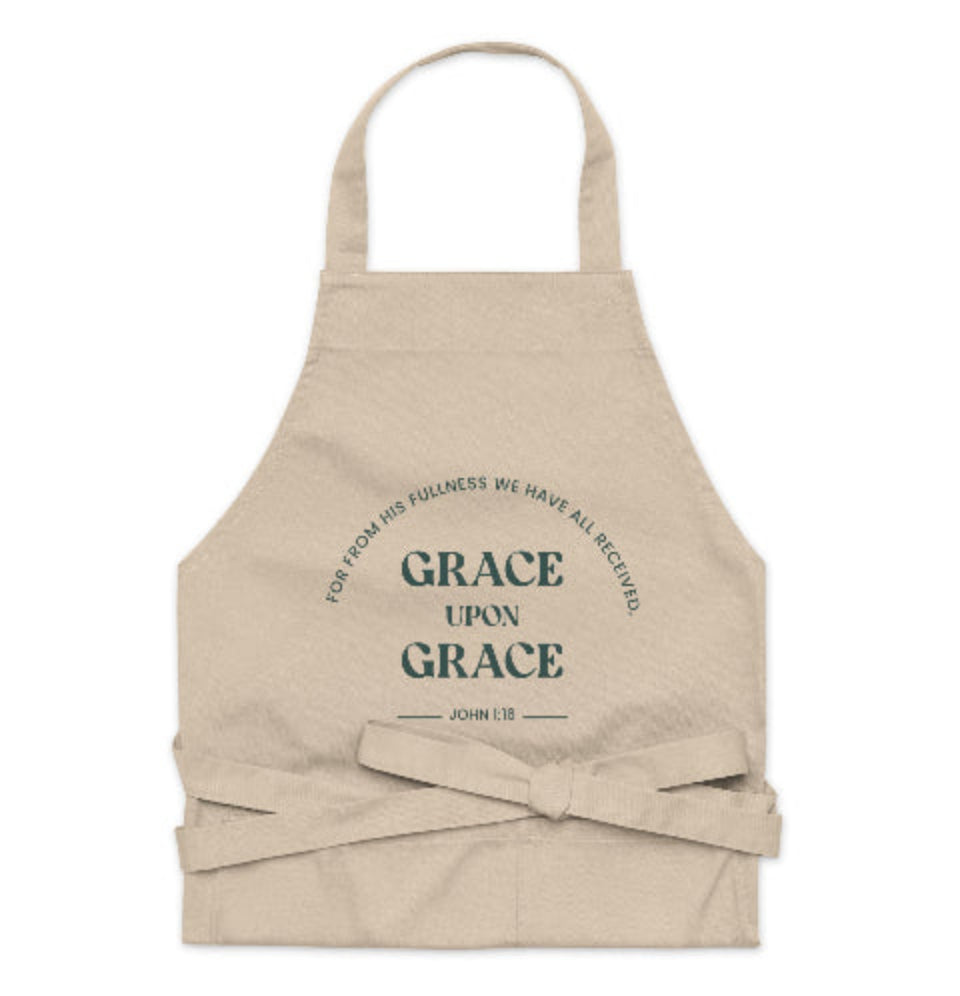 A beige organic cotton apron with a large front pocket, featuring the text 'GRACE UPON GRACE' from John 1:16 printed in a circular pattern.