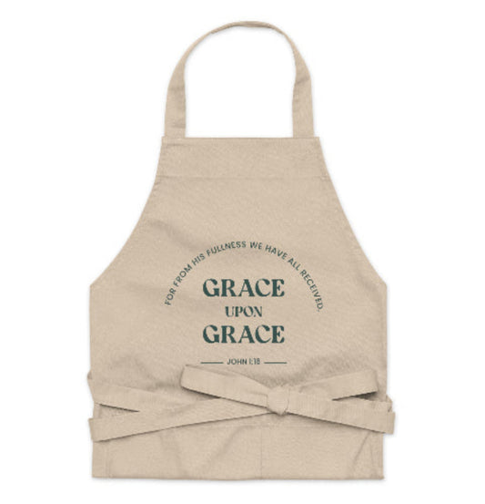 A beige organic cotton apron with a large front pocket, featuring the text 'GRACE UPON GRACE' from John 1:16 printed in a circular pattern.